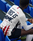 Person sitting in stadium seats wearing a white t-shirt with 'This is Home' text.