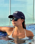 A woman in a swimming pool wearing a navy blue baseball cap with white 'WP' lettering, dark sunglasses, and a white bikini top.