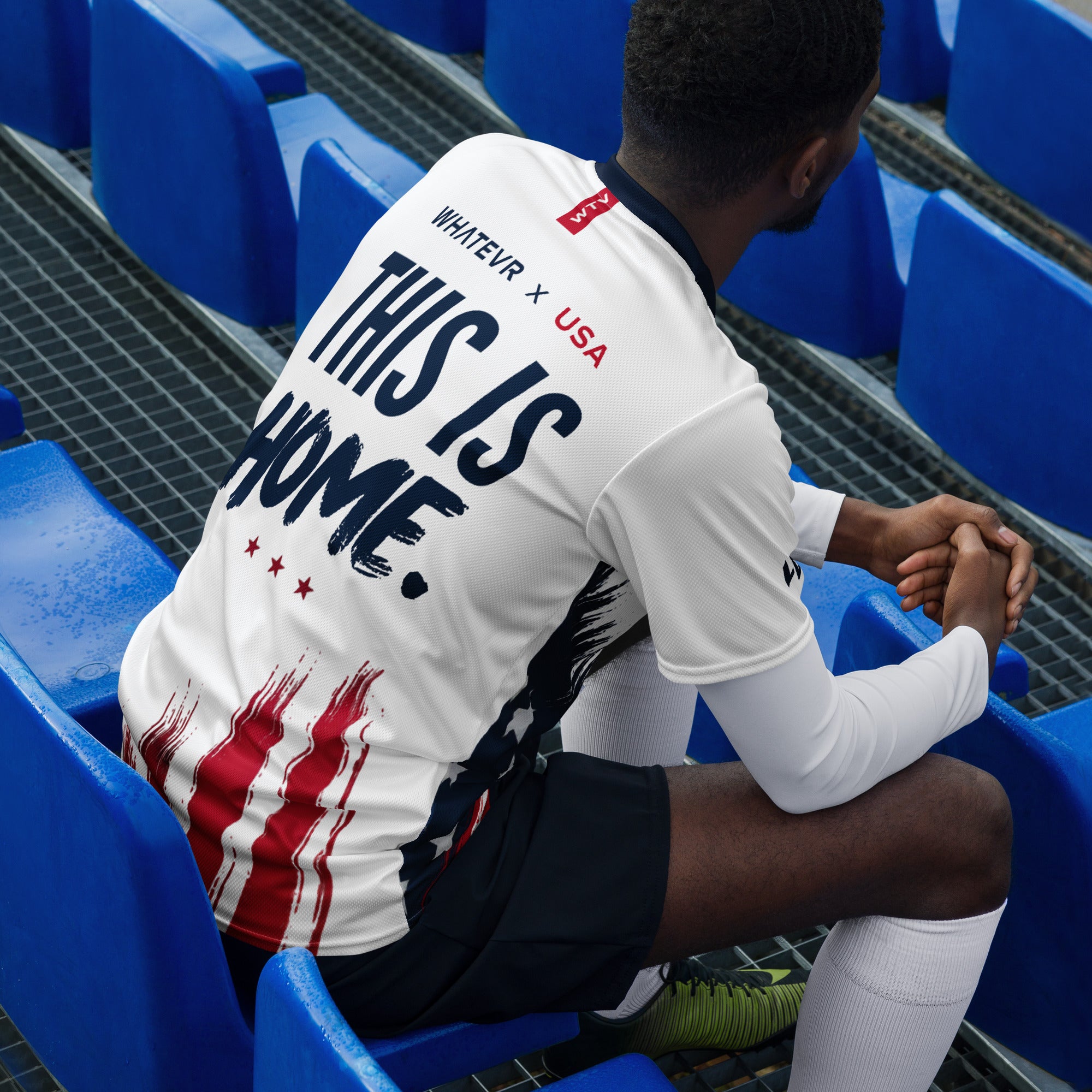 Person sitting in stadium seats wearing a white t-shirt with 'This is Home' text.